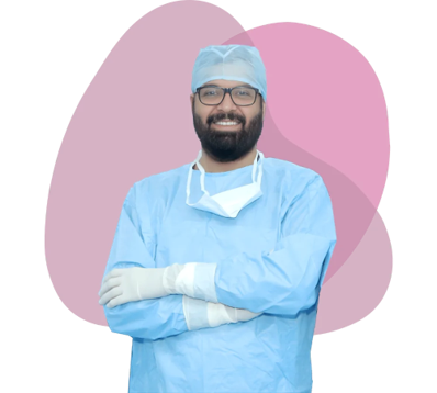 Smiling male surgeon in blue scrubs with a cap and mask worn under chin, arms crossed, standing against abstract pink shapes in the background